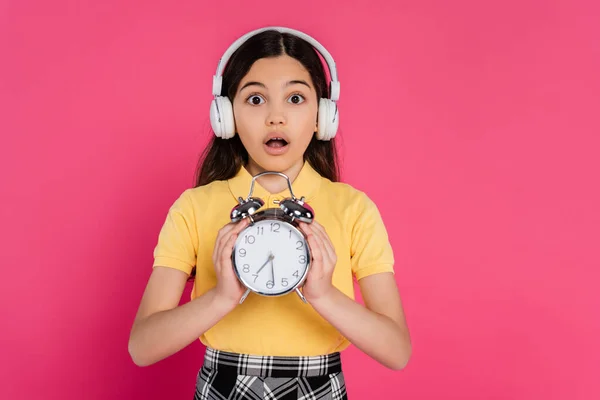Colegiala conmocionada en auriculares inalámbricos con reloj despertador vintage aislado en rosa, la vida estudiantil - foto de stock