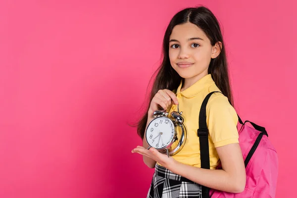 Feliz morena estudante de pé com mochila e segurando relógio de alarme vintage isolado em rosa — Fotografia de Stock
