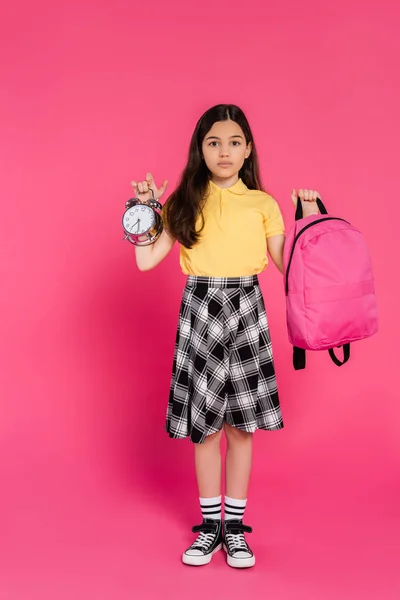 Full length, brunette schoolgirl standing with backpack and holding vintage alarm clock on pink — Stock Photo