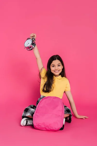Positive und brünette Schulmädchen sitzt mit Rucksack, hält Vintage-Wecker auf rosa — Stockfoto