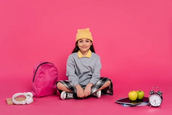Heureuse écolière en bonnet chapeau assis près du sac à dos, cahiers, écouteurs, pommes et réveil — Photo de stock