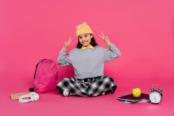 Happy schoolgirl in beanie hat showing v sign, notebooks, headphones, apple, backpack, alarm clock — Stock Photo