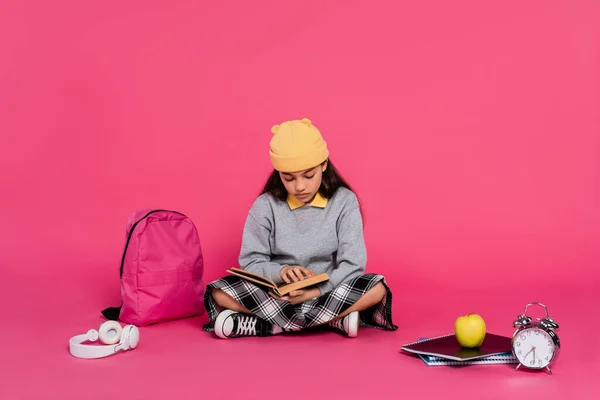 Schoolgirl in beanie hat reading book, sitting near headphones, apple, backpack, alarm clock — Stock Photo