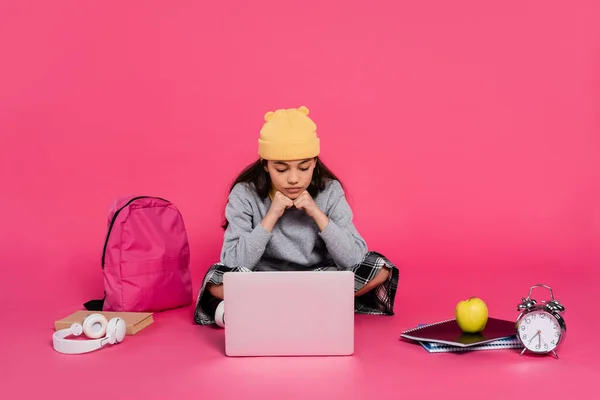 Estudante em chapéu gorro usando laptop, sentado perto de fones de ouvido, maçã verde, mochila, despertador — Fotografia de Stock