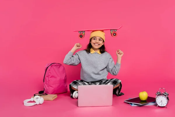 Chica feliz en gorro sombrero sentado con penny board en la cabeza, ordenador portátil, auriculares, manzana, despertador - foto de stock