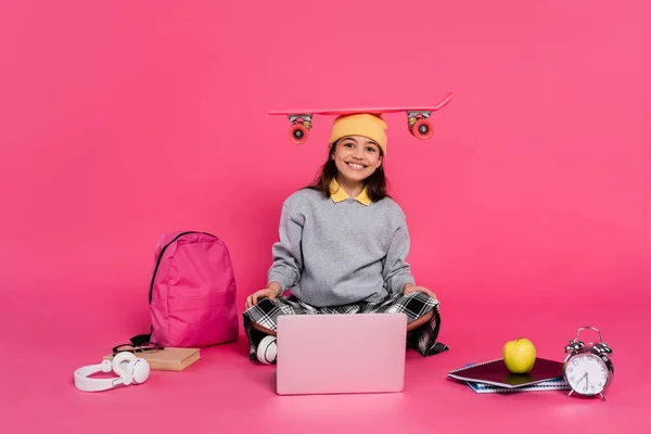 Sonrisa, chica en gorro sombrero sentado con penny board en la cabeza, ordenador portátil, auriculares, manzana, reloj despertador - foto de stock