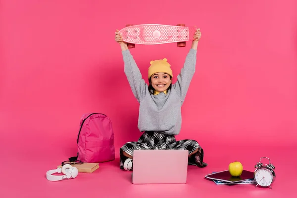 Excitado, chica en gorro sombrero celebración penny board en la cabeza, ordenador portátil, auriculares, manzana, despertador - foto de stock