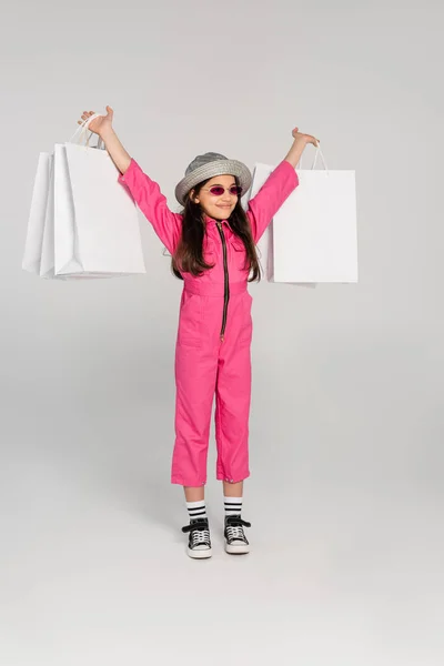 Chica excitada en elegante traje rosa y sombrero panama celebración de bolsas de compras sobre fondo gris - foto de stock