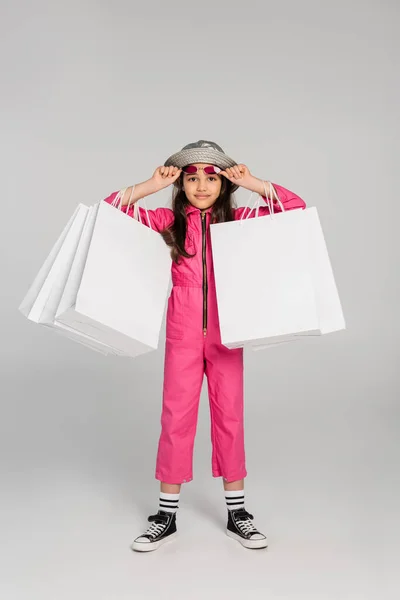 Chica en traje elegante y sombrero panama sosteniendo bolsas de compras en gris, ajustando gafas de sol de color rosa - foto de stock