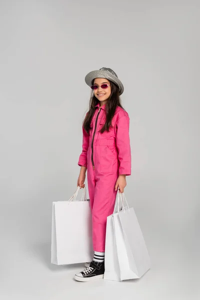 Chica alegre en traje elegante, gafas de sol y sombrero panama sosteniendo bolsas de compras sobre fondo gris - foto de stock