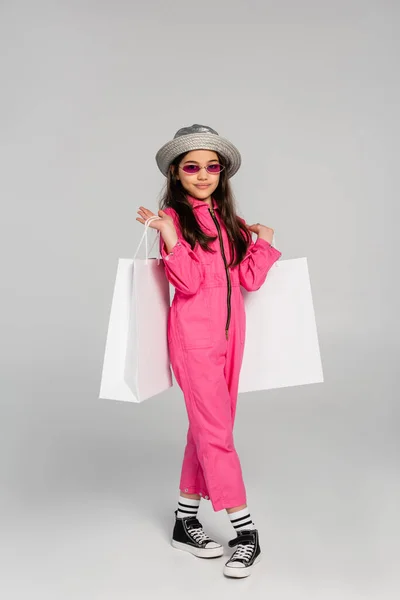 Chica sonriente en traje elegante, gafas de sol y sombrero panama sosteniendo bolsas de compras sobre fondo gris - foto de stock