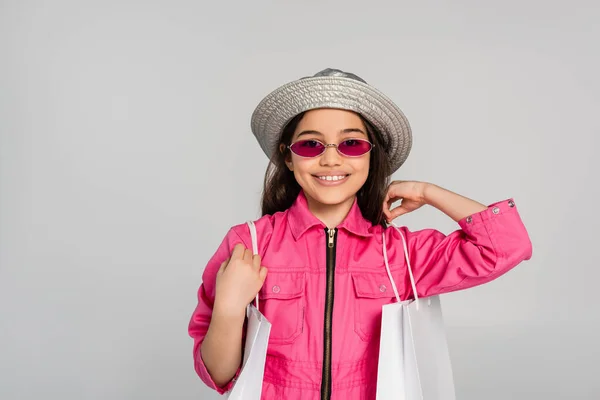 Chica sonriente en traje elegante, gafas de sol de color rosa y sombrero de panama posando con bolsas de compras en gris - foto de stock