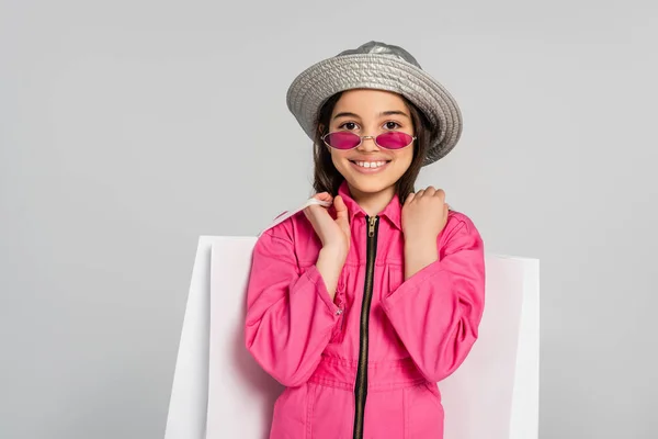 Fille positive en tenue élégante, lunettes de soleil roses et chapeau panama posant avec des sacs à provisions sur gris — Photo de stock