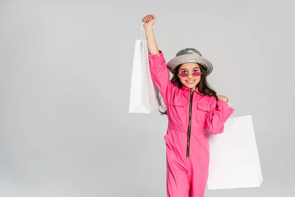 Положительная девушка в модном наряде, розовые солнцезащитные очки и панамская шляпа позируют с пакетами покупок на сером — стоковое фото