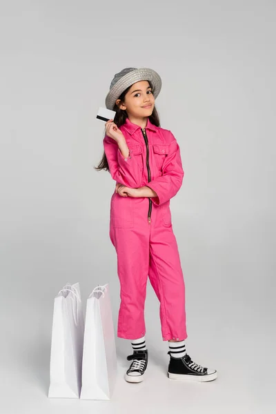 Glückliches Mädchen in trendigem Outfit und Panamahut posiert neben Einkaufstüten und hält Kreditkarte auf grau — Stockfoto