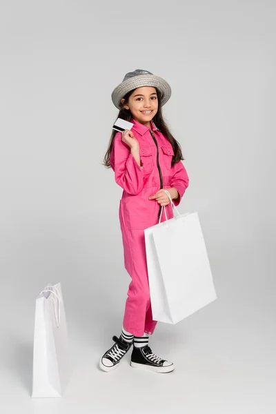 Chica sonriente en traje de moda y sombrero de panama posando cerca de bolsas de compras, la celebración de la tarjeta de crédito en gris - foto de stock