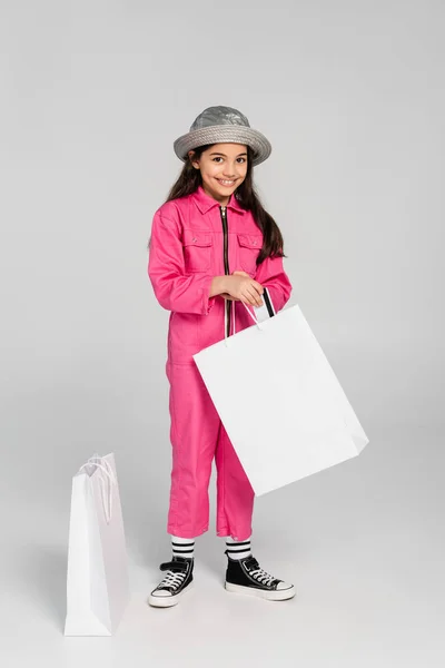 Chica feliz en traje de moda y sombrero de panama poner tarjeta de crédito en bolsas de compras, fondo gris - foto de stock