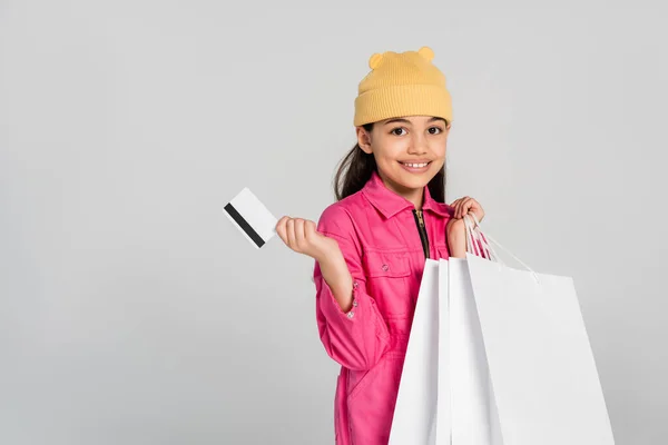 Glückliches Mädchen mit Beanie-Hut, Kreditkarte isoliert auf grauem, trendigem Outfit, Kind-Shopping, positiv — Stockfoto
