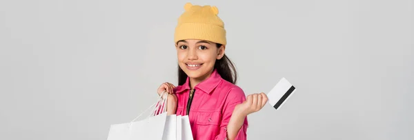 Fille heureuse en bonnet chapeau tenant la carte de crédit isolée sur gris, tenue à la mode, achats pour enfants, bannière — Photo de stock