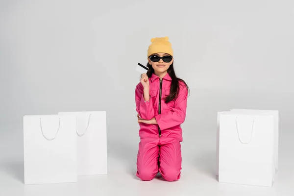 Menina feliz em chapéu gorro e óculos de sol segurando cartão de crédito em cinza, compras de crianças, roupa da moda — Fotografia de Stock