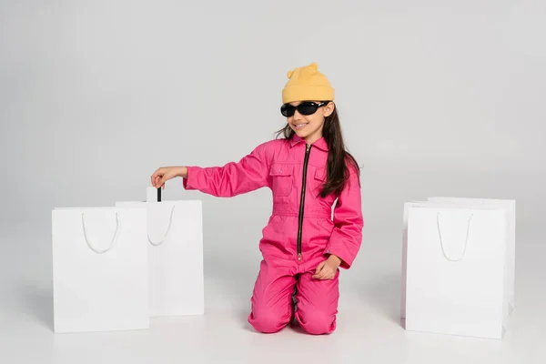 Allegra ragazza in cappello beanie e occhiali da sole con carta di credito su grigio, shopping per bambini, vestito alla moda — Foto stock