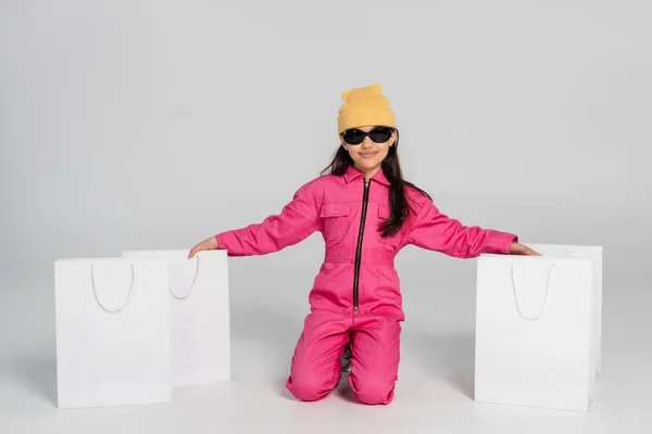 Alegre chica en gorro sombrero y gafas de sol sentado cerca de bolsas de compras en gris, niño, traje de moda - foto de stock