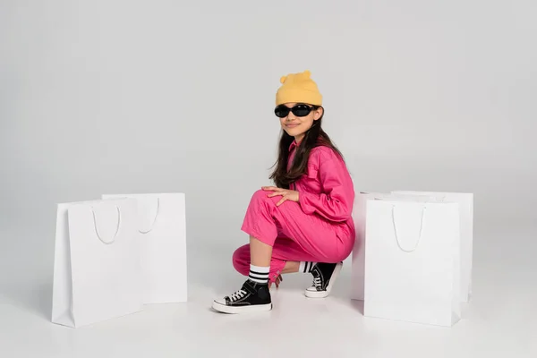 Ragazza alla moda in cappello beanie e occhiali da sole seduto in posa vicino a borse della spesa su grigio, bambino felice — Foto stock