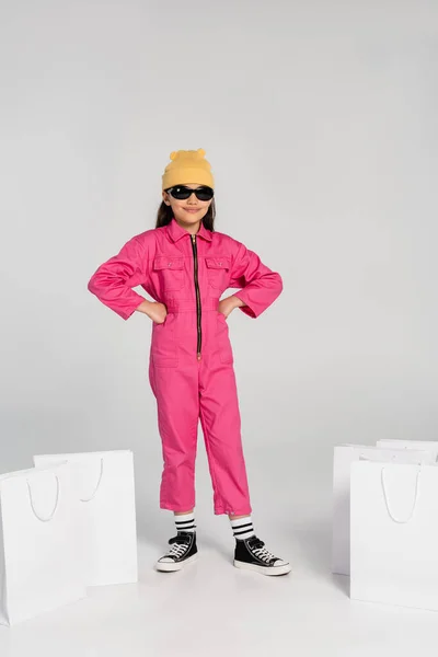 Chica de moda en gorro sombrero y gafas de sol sentado posando con las manos en las caderas cerca de las bolsas de compras - foto de stock