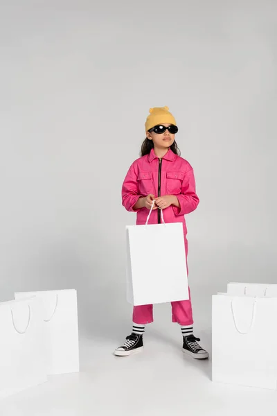 Stylish preteen girl in beanie hat and sunglasses standing and holding shopping bags on grey — Stock Photo