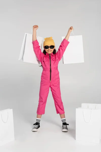Emocionado niña preadolescente en gorro sombrero y gafas de sol sosteniendo bolsas de compras sobre fondo gris, estilo - foto de stock