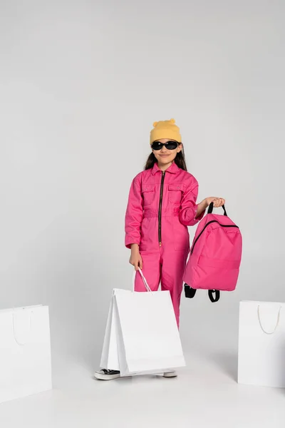 Happy schoolgirl in beanie hat and sunglasses holding shopping bags and backpack on grey, style — Stock Photo