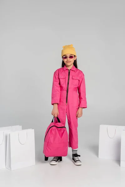 De vuelta a la escuela, chica feliz en gorro y gafas de sol sosteniendo bolsas de compras y mochila en gris - foto de stock