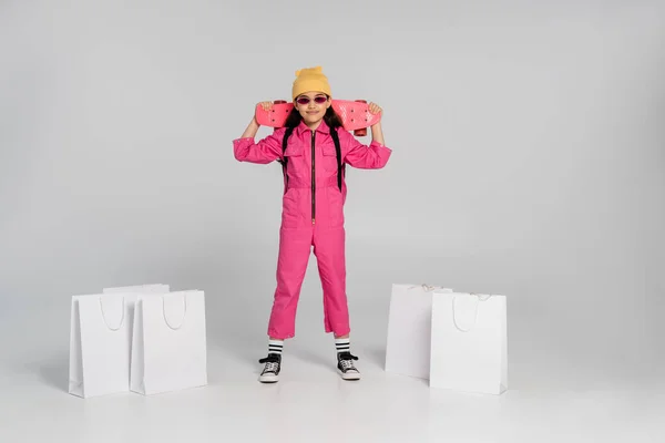 Menina feliz em gorro e óculos de sol elegantes segurando penny board, sacos de compras em fundo cinza — Fotografia de Stock