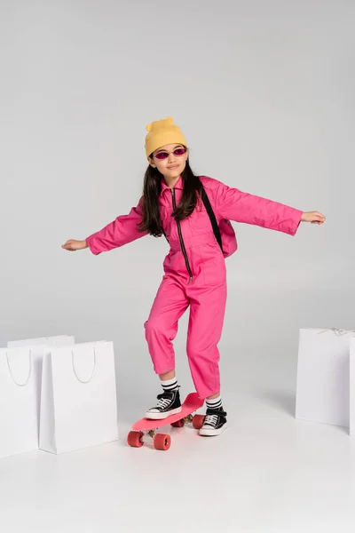 Chica alegre en gorro y gafas de sol con estilo a caballo penny board, bolsas de compras sobre fondo gris - foto de stock