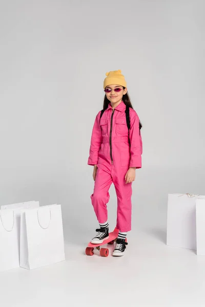 Chica positiva en gorro y gafas de sol de color rosa a caballo penny board, bolsas de compras sobre fondo gris - foto de stock