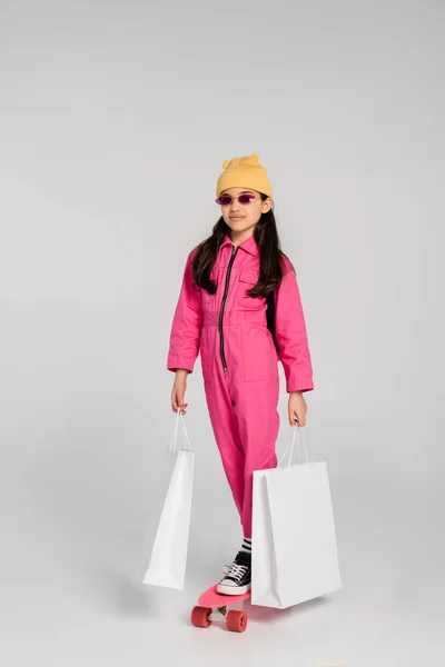 Chica con estilo en gorro y gafas de sol de color rosa a caballo penny board y la celebración de bolsas de compras en gris - foto de stock