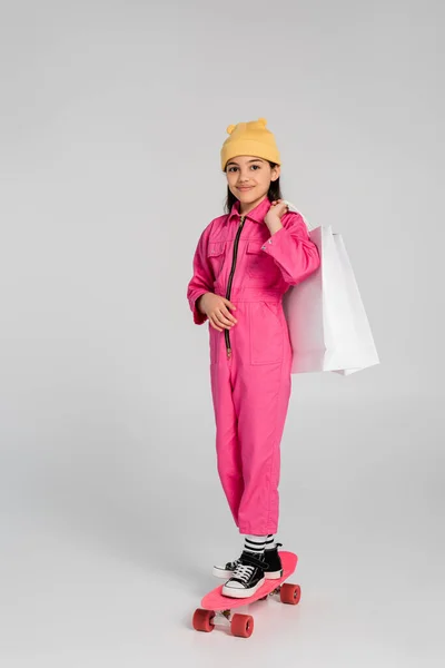 Glückliches Mädchen mit Mütze und rosa Outfit reitet Penny Board und hält Einkaufstüten auf grau — Stockfoto