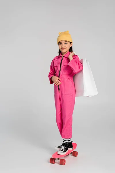 Happy girl in beanie hat and pink outfit riding penny board and holding shopping bags on grey — Stock Photo