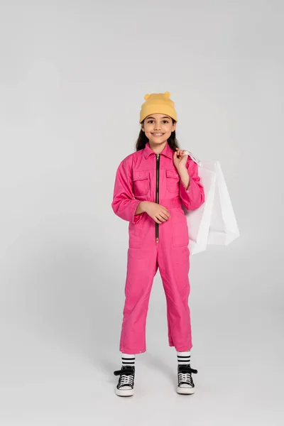 Chica feliz en gorro amarillo sombrero y traje rosa sosteniendo bolsas de compras y de pie en gris, moda - foto de stock