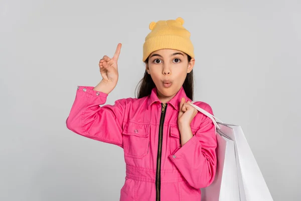 Ragazza stupita in cappello beanie e vestito rosa con borse della spesa e puntando verso l'alto, sfondo grigio — Foto stock