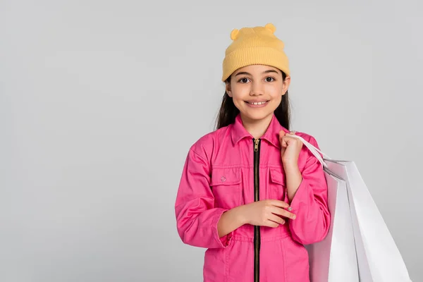 Allegra ragazza in cappello beanie e vestito rosa con borse della spesa e puntamento verso l'alto, sfondo grigio — Foto stock