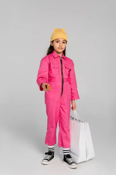 Chica con estilo en gorro sombrero dando tarjeta de crédito y bolsas de compras en gris, traje de moda, compras para niños - foto de stock