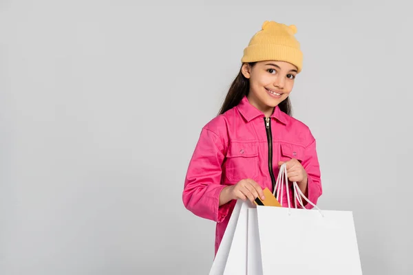 Happy girl in beanie hat putting credit card into shopping bag on grey, trendy outfit, kid shopping — Stock Photo