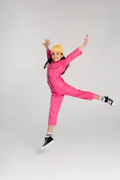 Happy girl in beanie hat and stylish pink outfit jumping with outstretched hands on grey, have fun — Stock Photo