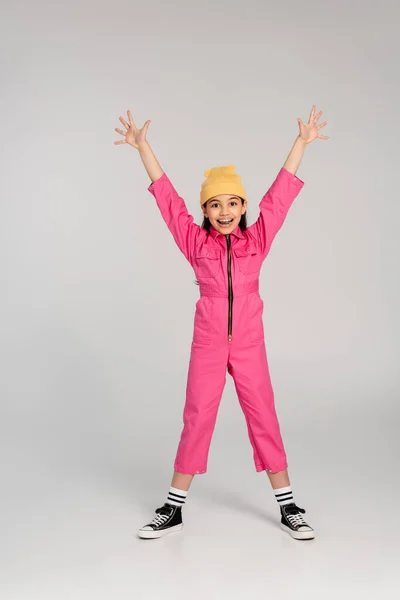 Happy girl in beanie hat and pink outfit standing with outstretched hands on grey, have fun, style — Stock Photo