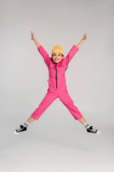 Excited girl in trendy beanie hat and pink outfit jumping with outstretched hands on grey, have fun — Stock Photo