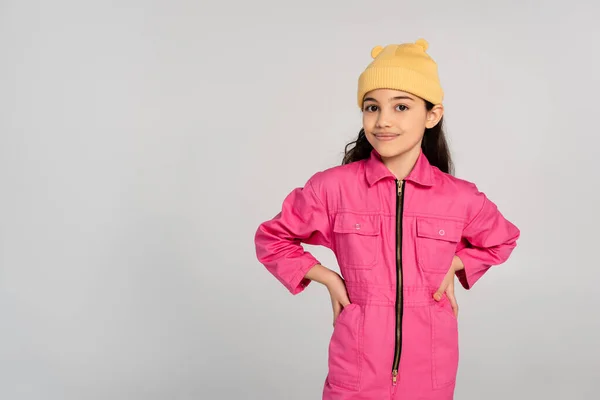 Heureux enfant en bonnet jaune chapeau et tenue rose debout avec les mains sur les hanches isolé sur gris, mode — Photo de stock