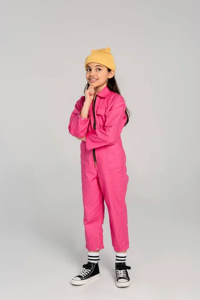 Happy kid in beanie hat and pink outfit standing and looking away, thinking on grey backdrop — Stock Photo