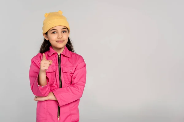 Bambino felice in cappello beanie giallo e vestito rosa che punta alla fotocamera su sfondo grigio, bambino alla moda — Foto stock