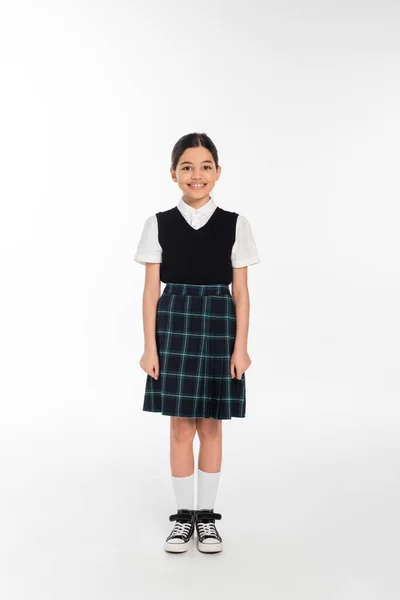 Comprimento total, estudante feliz em uniforme de pé e olhando para a câmera no branco, menina na saia — Fotografia de Stock